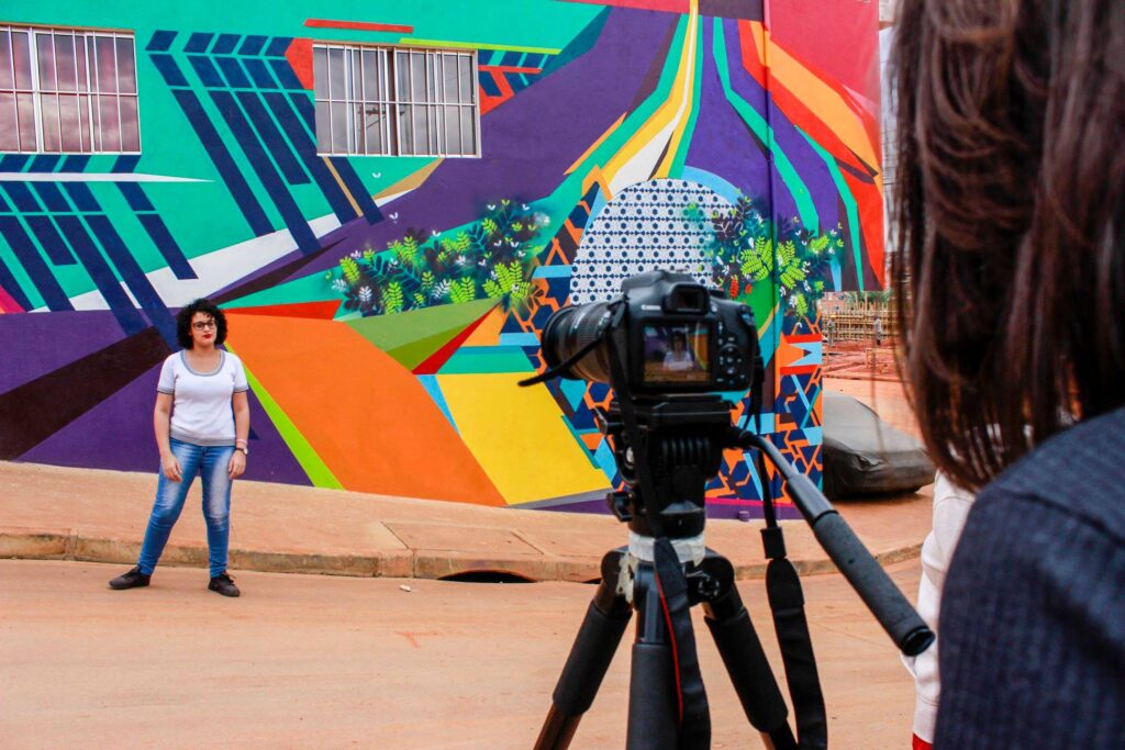 Uma mulher posa para uma foto em frente a um mural colorido, enquanto uma câmera em um tripé a captura.
