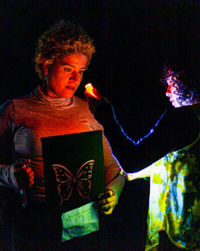 Duas pessoas em ambiente escuro, uma com livro de borboleta, outra iluminando com luz.